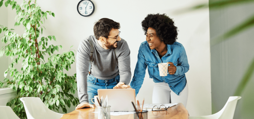 A white man and black woman stand over a table laughing as the man gestures toward a laptop device on the table. The woman is holding a coffee mug.