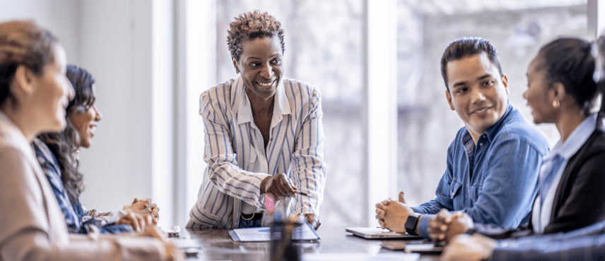 A black woman smiles while standing at the head of a conference table where a group is sitting. The woman and the group are focused on another woman at the table who is speaking.